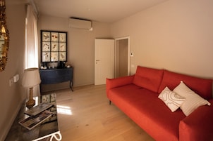 Una sala de estar con un sofá rojo, una mesa de centro con revistas, una lámpara y una composición botánica enmarcada en la pared.