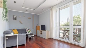 Ein modernes Wohnzimmer mit einem grauen Sofa, Holzboden, einem Fernseher auf einem weißen Schrank und einem Balkon mit Blick.