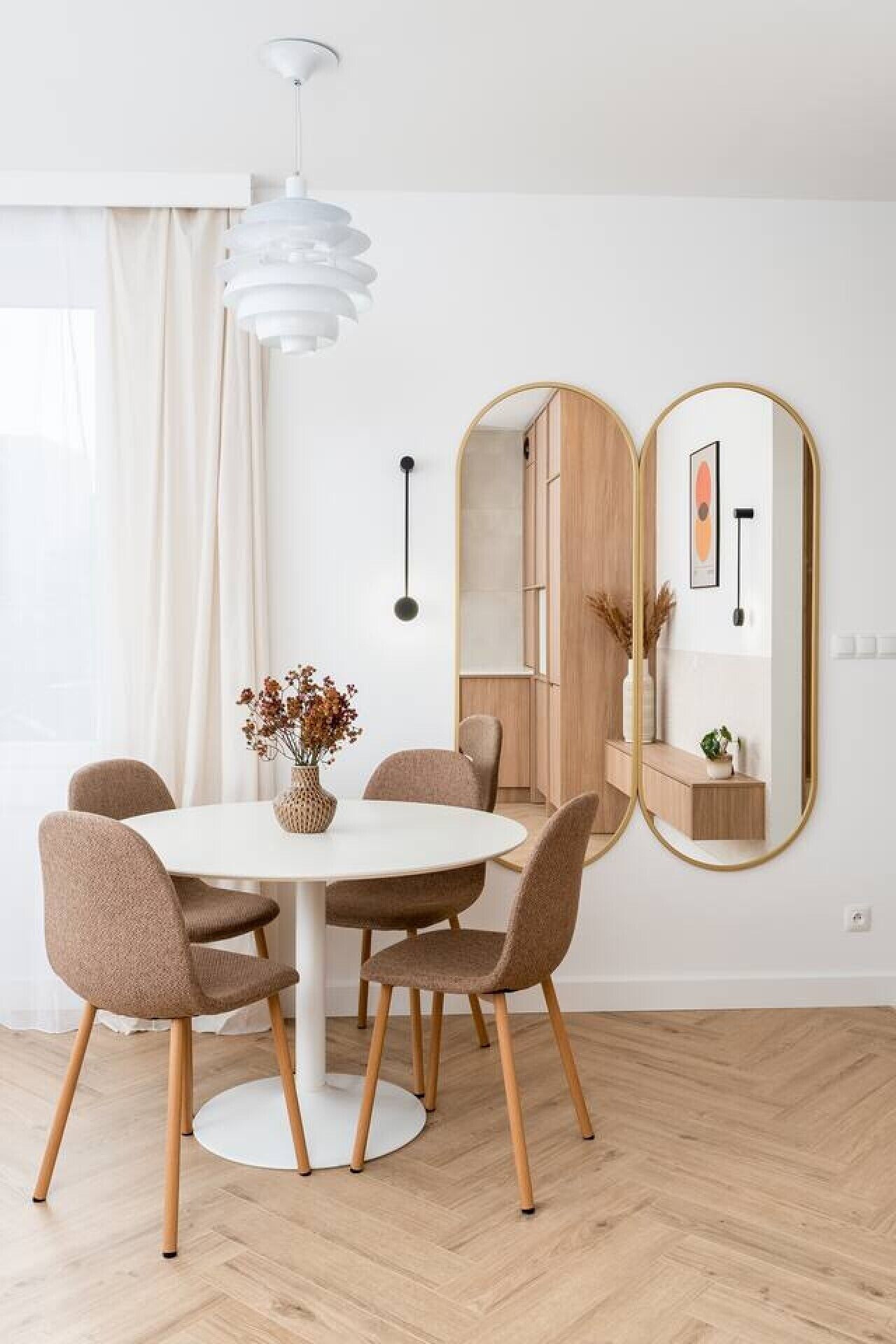 Un espace repas moderne avec une table ronde, six chaises, un vase contenant des fleurs séchées et un grand miroir au mur.