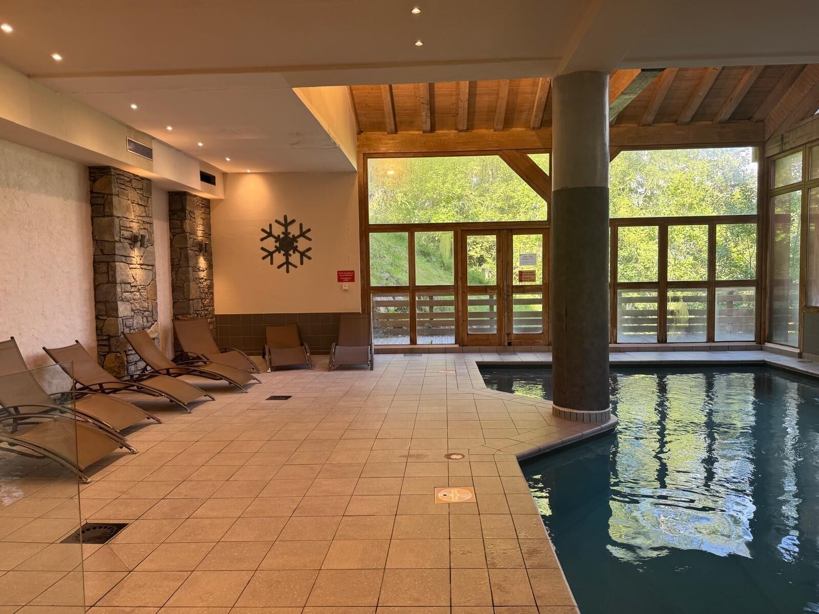 An indoor swimming pool with tiled flooring, lounge chairs, a stone wall, and large windows.