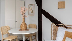 Een kamer met een ronde tafel, rieten stoelen, een witte kast, een bed met een geweven hoofdeinde, en een wandlamp.