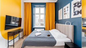 Habitación de hotel moderna con cama, televisión, ventana con cortinas y una obra de arte en la pared.