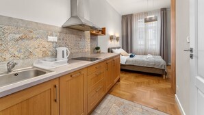 Eine kleine Küche mit Spüle, Herd und Holzschränken. Im Hintergrund ist ein Bett mit grauer Decke und ein Fenster mit Vorhängen zu sehen.