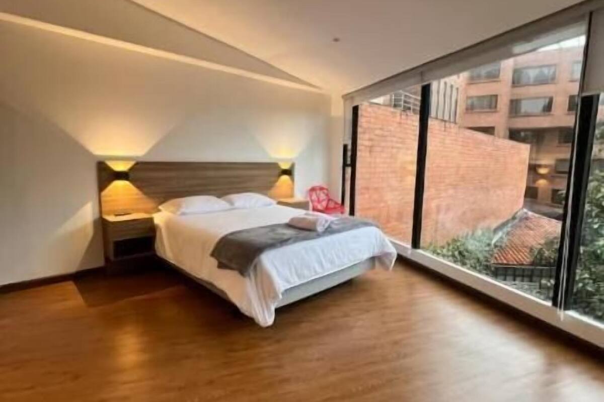 A modern bedroom with a large bed, bedside lamps, and a view of a building through large windows.