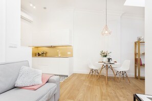 Una sala moderna con un sofá gris, una mesa de comedor con sillas blancas y una zona de cocina con armarios blancos y encimera de madera.