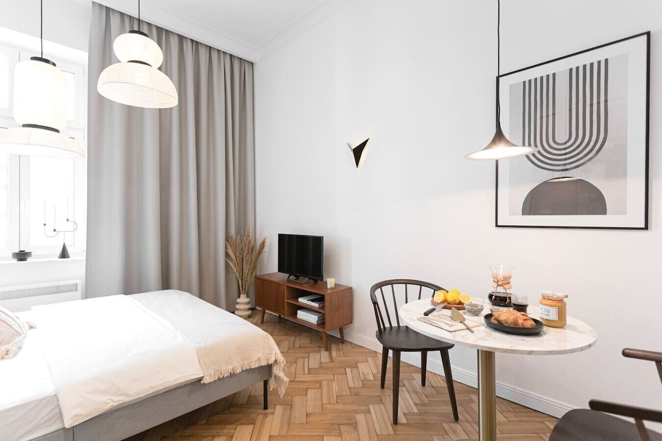 Une chambre d’hôtel moderne avec un lit, un téléviseur posé sur un meuble en bois, une table à manger avec des éléments pour le petit-déjeuner et une grande fenêtre avec des rideaux.