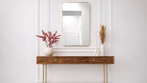 Eine weiße Wand mit einem großen Spiegel, ein Holzkonsole mit goldenen Füßen und zwei Vasen mit roten Blumen und getrockneten Pflanzen.