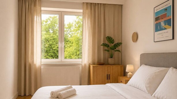 Een hotelkamer met een groot bed, een raam met gordijnen, een nachtkastje met een lamp en een lijst met een foto aan de muur.