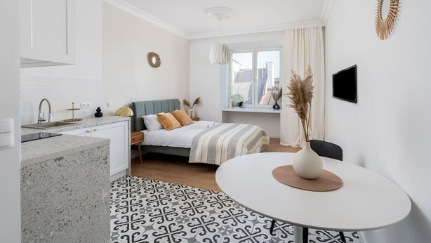 Una habitación de hotel moderna con cocina, una cama con colcha a rayas, una mesa de comedor redonda y un ventanal con vistas.