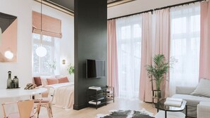 En moderne stue med et fladskærms-tv, et spiseområde med bord og stole, samt et stort vindue med gardiner.