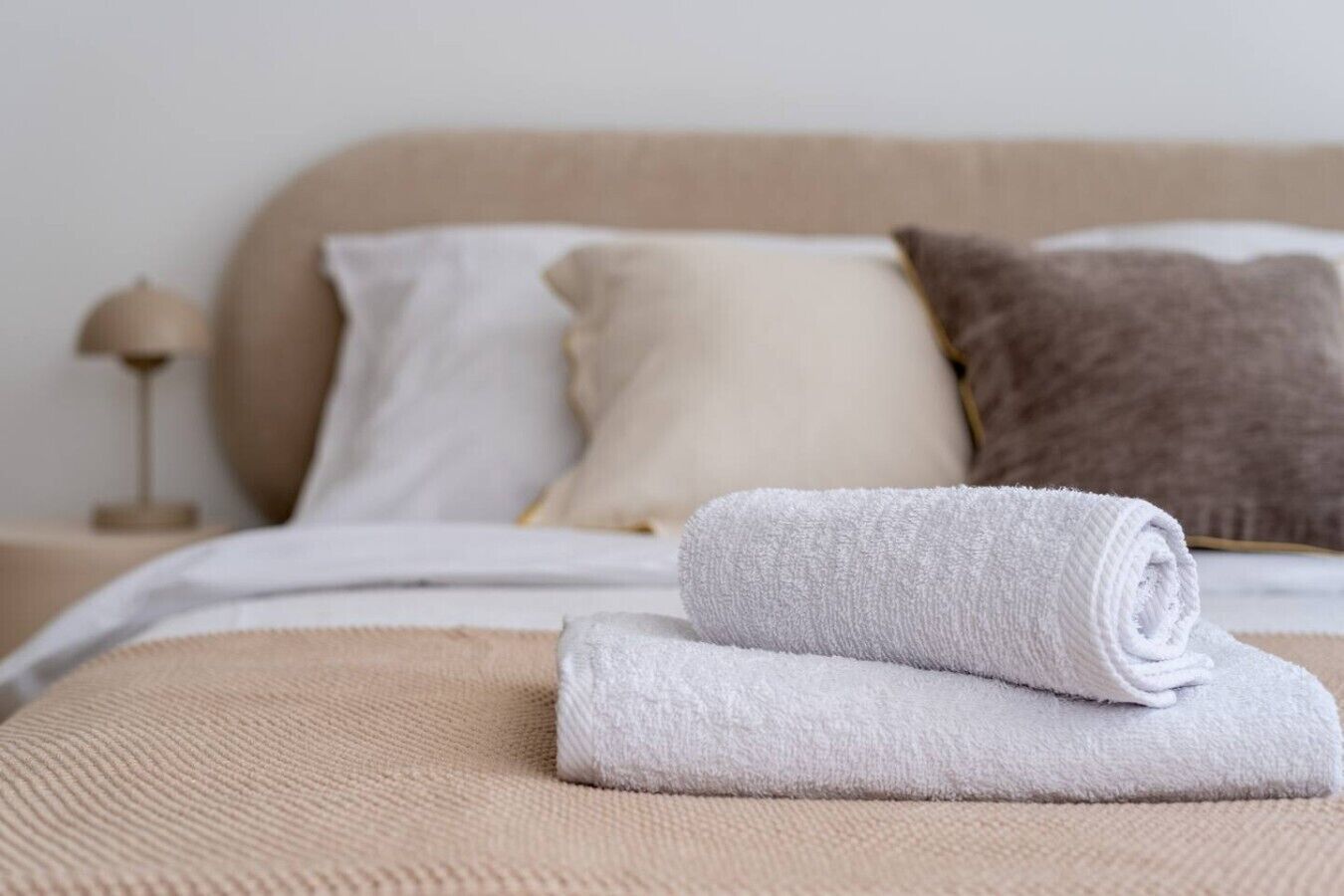 A neatly made bed with a rolled white towel, a beige headboard, and a bedside lamp.