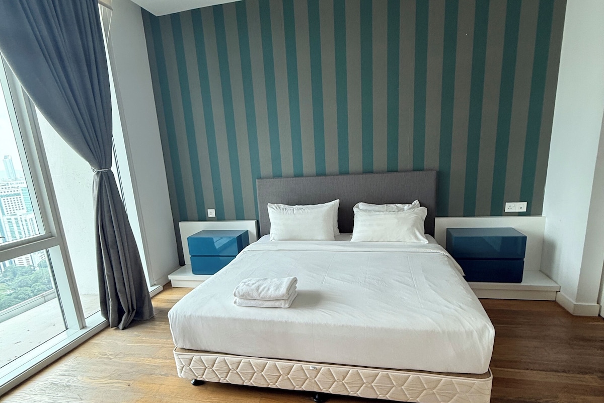 Un grand lit avec un matelas matelassé, deux oreillers et une serviette pliée posée sur le lit. Il y a deux tables de chevet avec des boîtes de rangement. La pièce est dotée d’un mur à rayures et d’une grande fenêtre avec des rideaux.