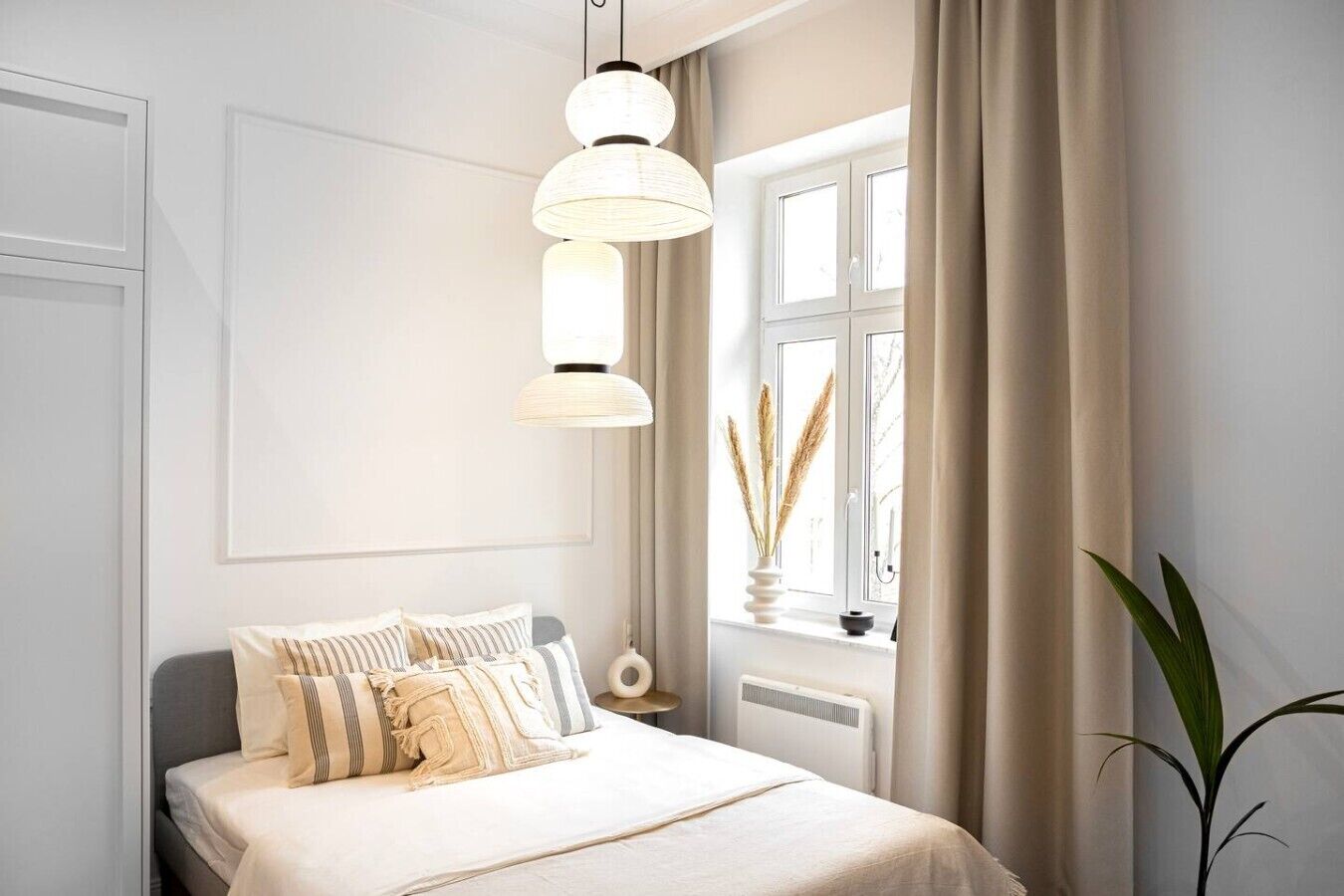 A bedroom with a bed, a hanging light fixture, a window with curtains, and a plant.