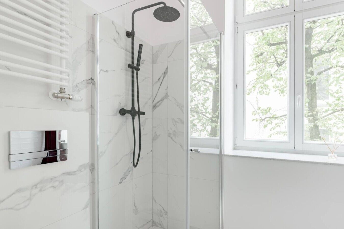 A modern bathroom with a glass shower enclosure, a wall-mounted mirror, and a window with a view of trees.