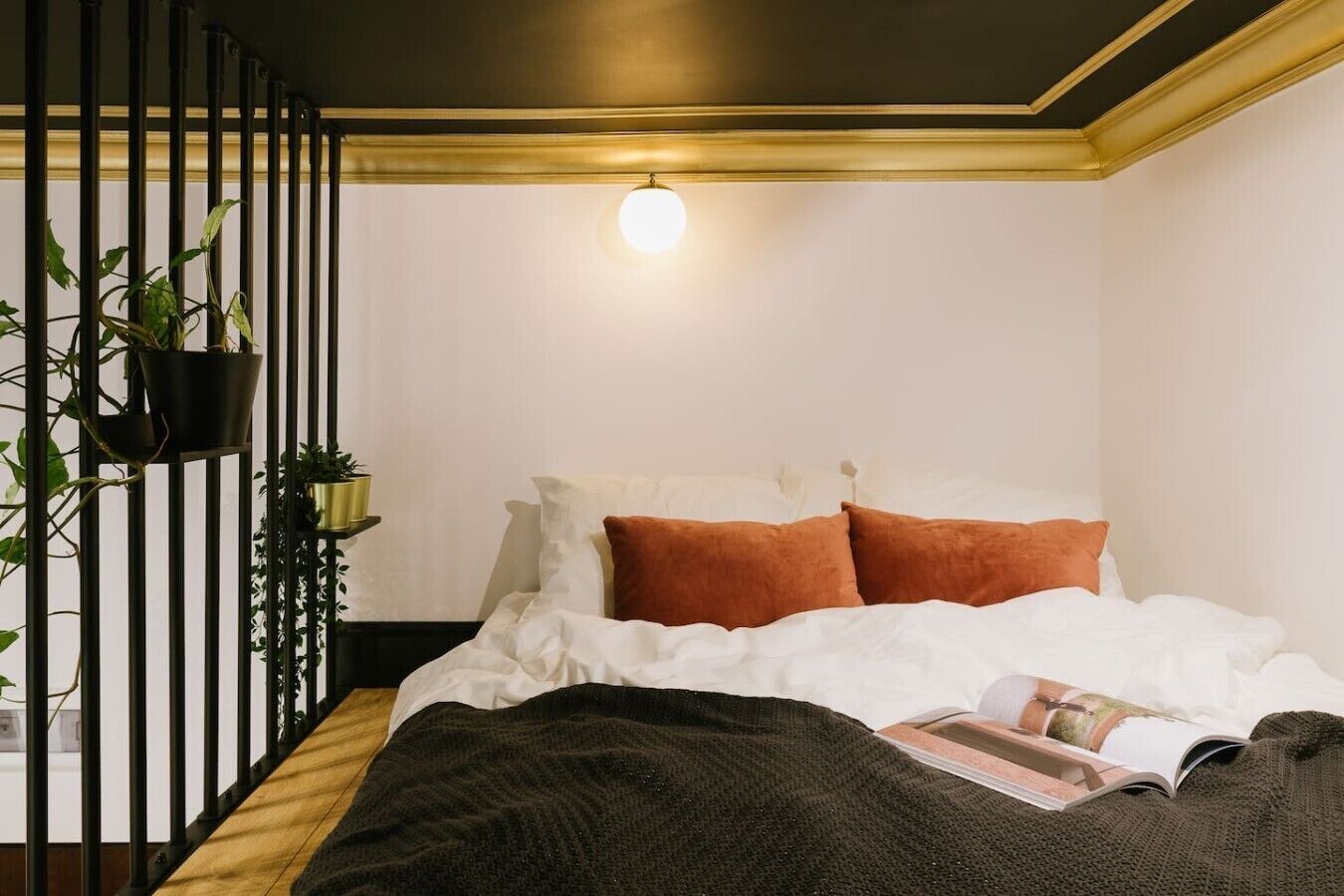 A bedroom with a bed, pillows, a blanket, a magazine, and potted plants.
