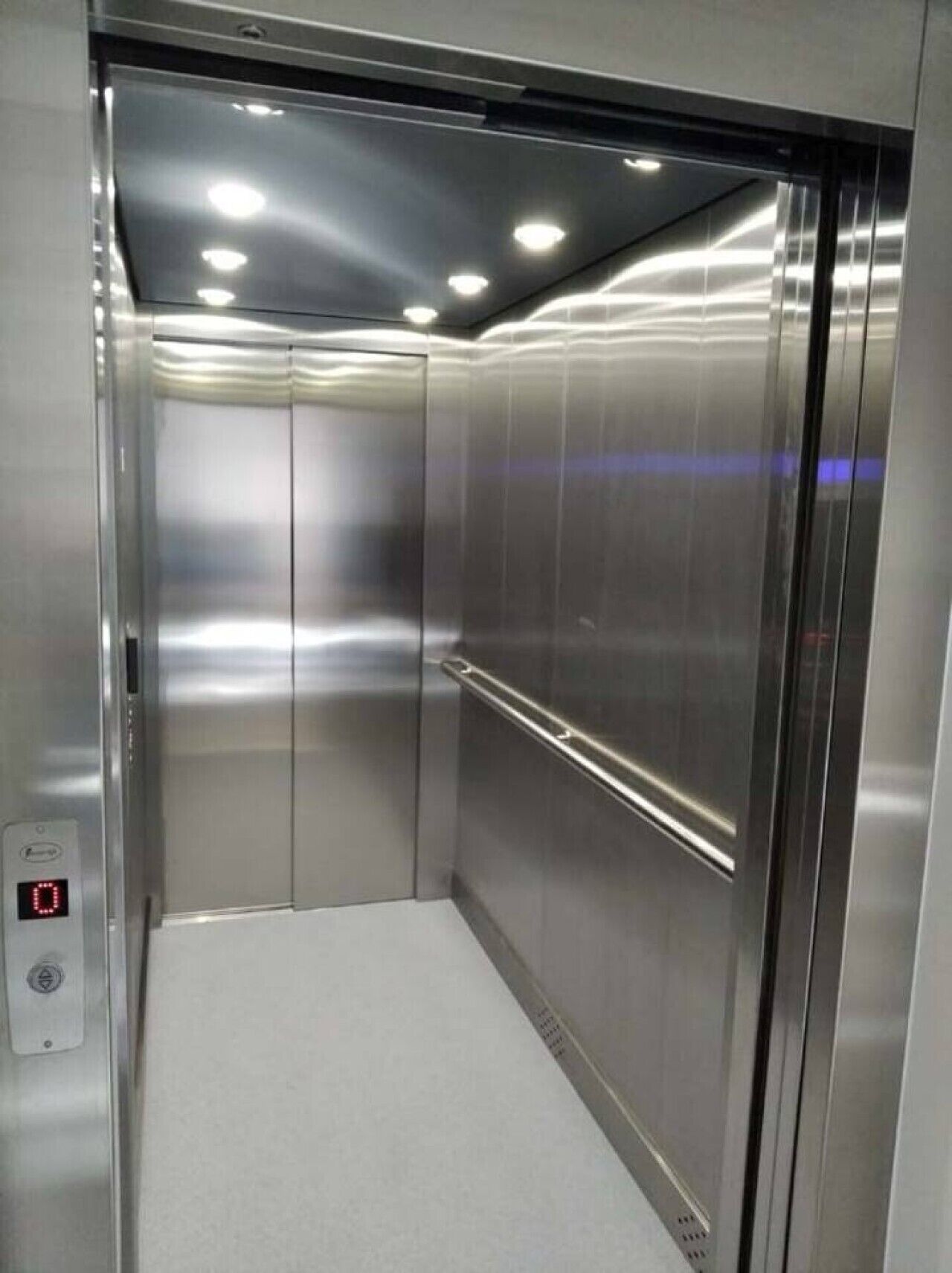 Interior de elevador metálico com corrimão, painel de comandos e iluminação no teto.