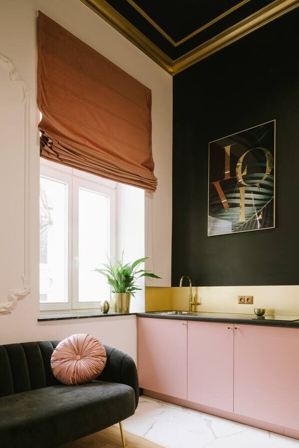 Eine moderne Küche mit schwarzem Fliesenspiegel, einem Fenster mit brauner Gardine, einer schwarzen Couch mit rosanen Kissen und einem Waschbecken mit goldfarbenen Armaturen.