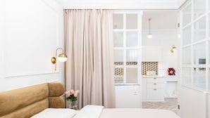 Un dormitorio con cama, mesita de noche, un florero con flores, ventana con cortinas y vistas al baño y la cocina.