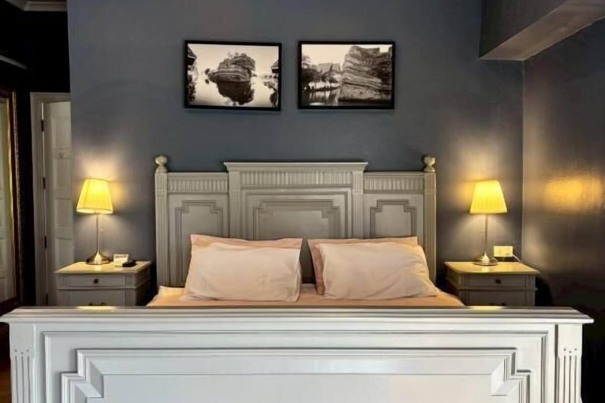 Una cama con cabecera blanca, dos lámparas de noche y dos cuadros en la pared.