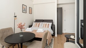 Una habitación de hotel moderna con una cama, una mesa redonda, una silla, un florero con flores y un cuadro en la pared.