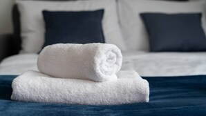 En pænt redt seng med hvide sengetæpper og mørke puder, et sammenrullet hvidt håndklæde ovenpå, og et mørkeblåt dynebetræk.