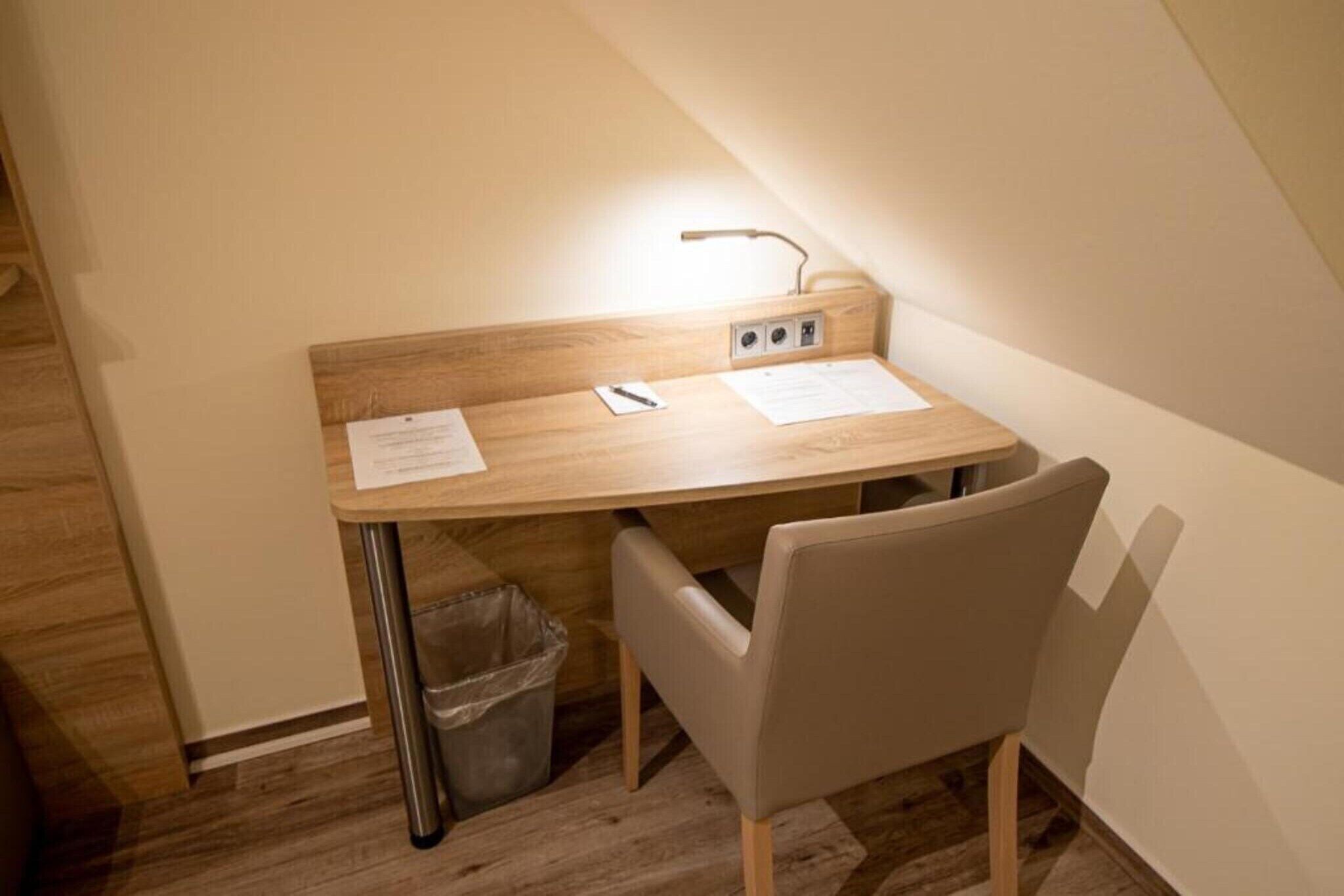 Une petite pièce bien éclairée, avec un bureau en bois, une chaise et une poubelle.