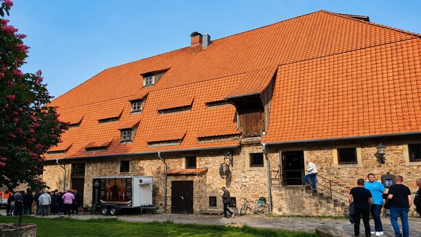 Un bâtiment en pierre avec un toit en tuiles, un escalier et un groupe de personnes à l’extérieur.