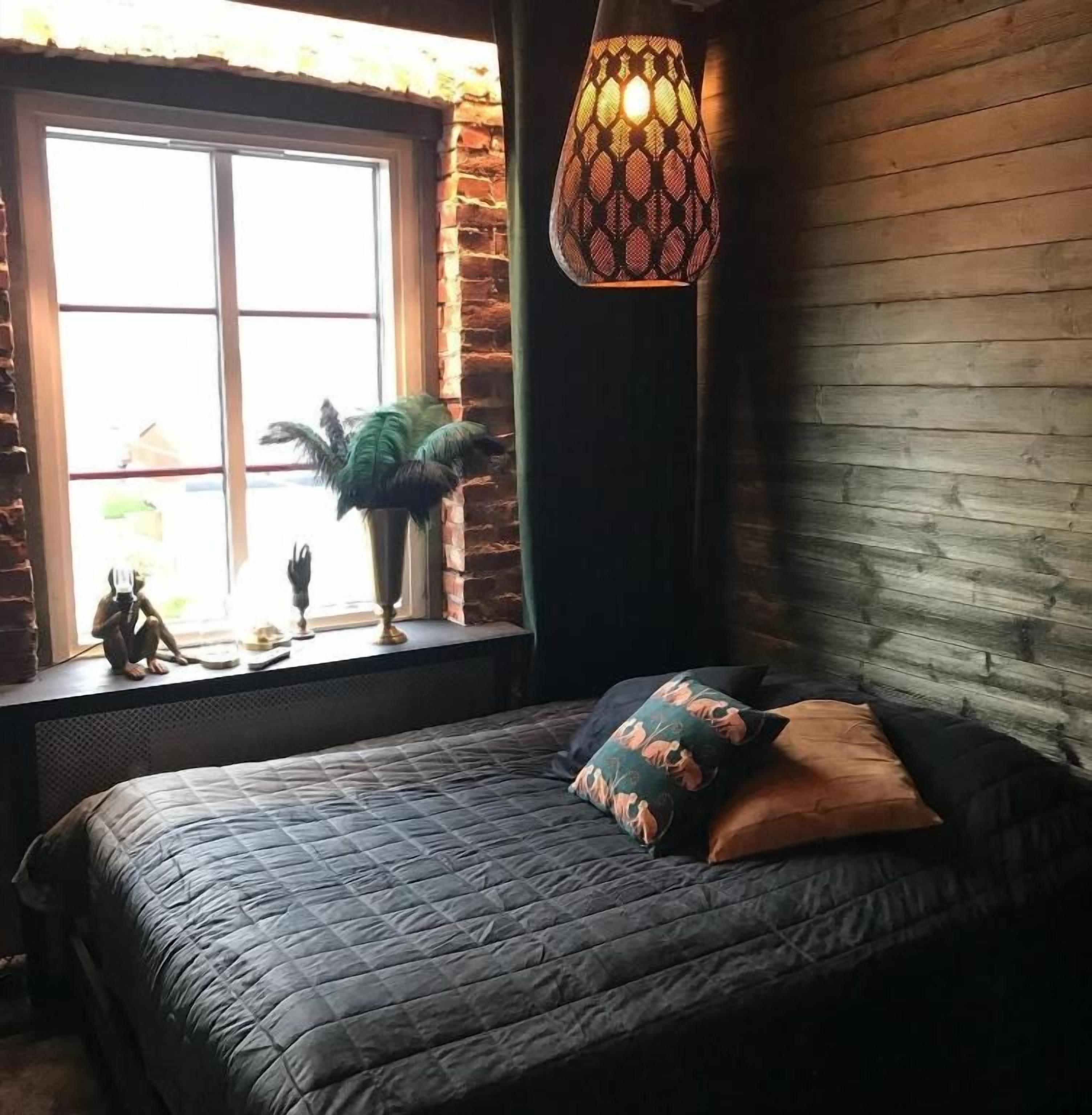 Un dormitorio con un ventanal grande, una cama con muchos almohadones, una lámpara colgante y una planta decorativa en el alféizar.