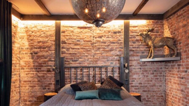 Un dormitorio con pared de ladrillo, cama con cabecera, lámpara colgante y escultura de animal montada.