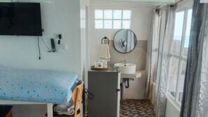 A small room with a bed, a desk, a refrigerator, a sink, and a window with curtains.