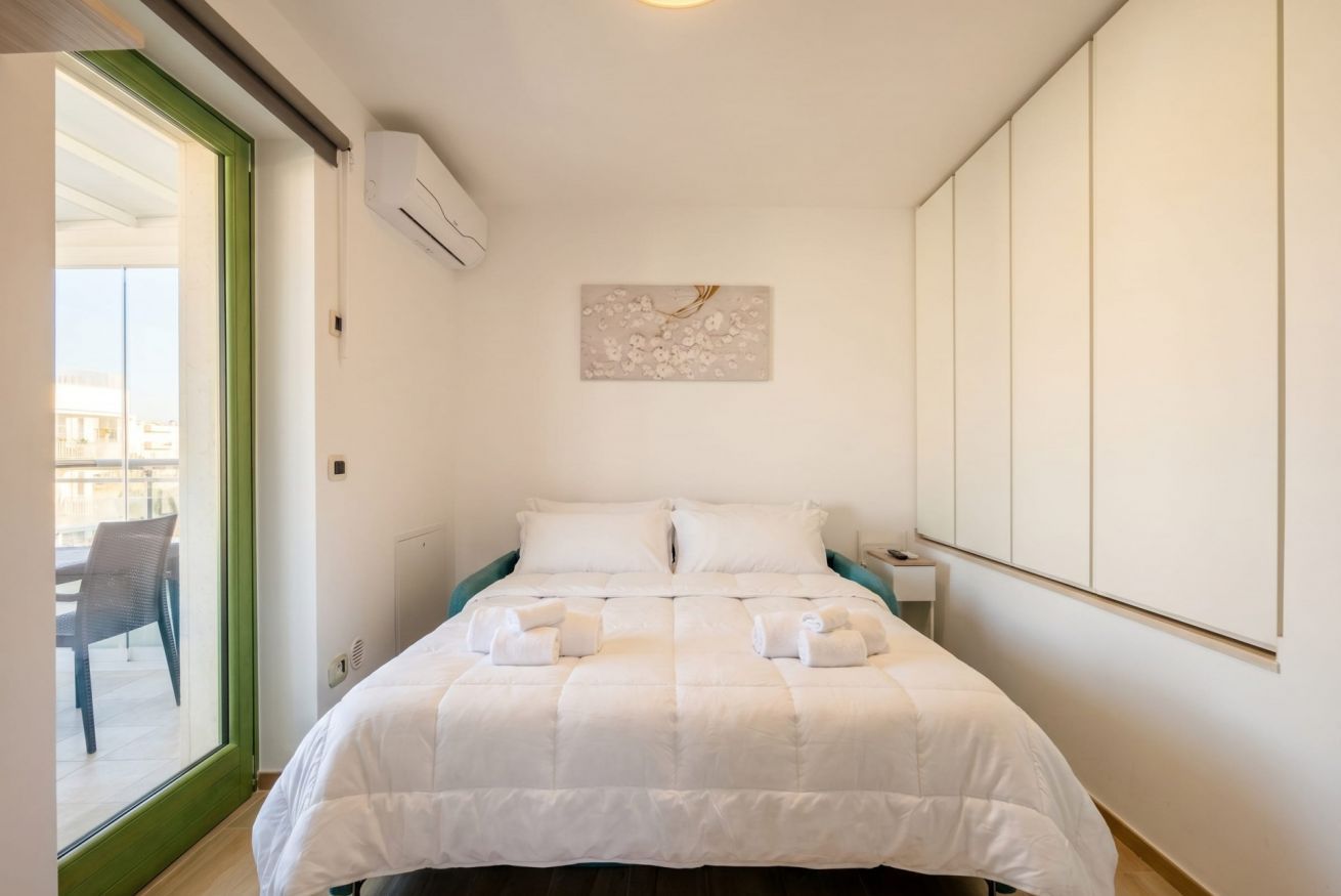 Une chambre d’hôtel avec un lit, une fenêtre encadrée de vert, un appareil de climatisation, une table de chevet et une œuvre d’art accrochée au mur.