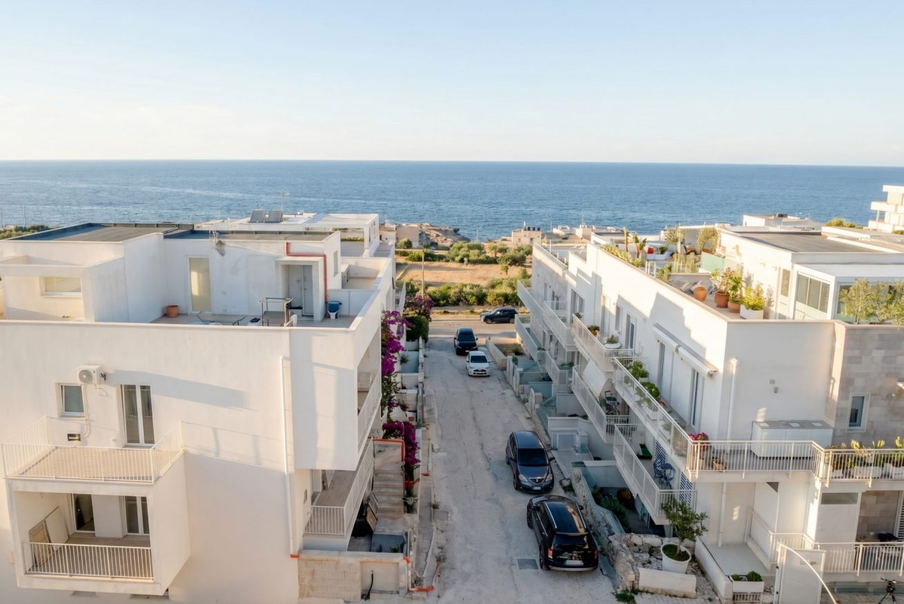 Un quartier résidentiel avec des bâtiments blancs, une rue étroite, des voitures garées et une vue sur la mer.