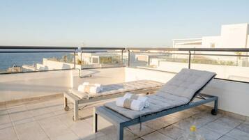 A rooftop terrace with two sun loungers, a glass table with a tray, and a clear view of the sea.