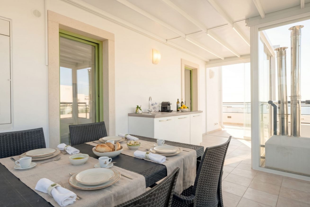 Un coin repas avec une table dressée pour quatre personnes, un comptoir de cuisine avec une machine à café, et une vue sur la mer grâce à de grandes fenêtres.
