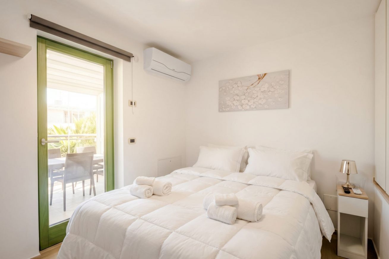 A hotel room with a bed, a green-framed door, an air conditioning unit, a small bedside table with a lamp, and a wall-mounted artwork.