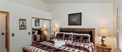 A neatly made bed with a patterned bedspread, a headboard, two bedside lamps, a framed picture above the bed, and a mirror on the wall.