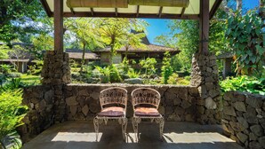 Une terrasse couverte avec deux chaises décoratives, un mur de pierre et une vue sur des bâtiments traditionnels et une végétation luxuriante.