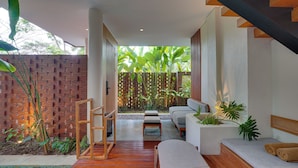 Un interior moderno con una escalera de madera, una zona de estar con cojines y vistas a un jardín con plantas y un muro de ladrillo.