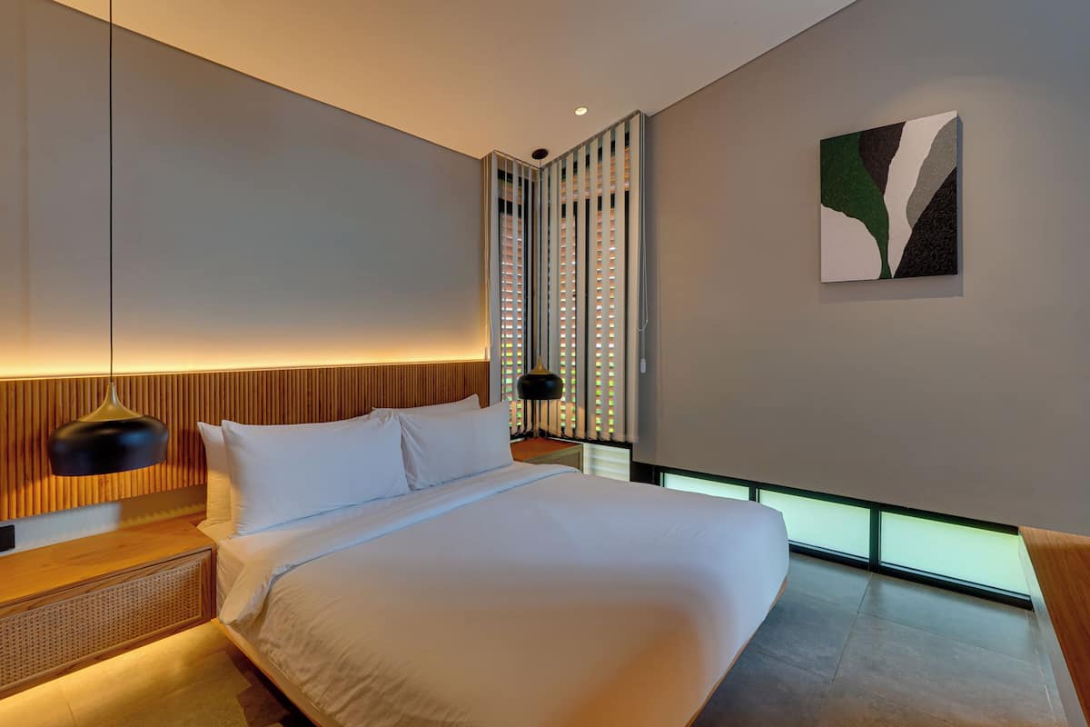 A modern hotel room with a large bed, a bedside table, a lamp, a wall-mounted artwork, and a window with a view.