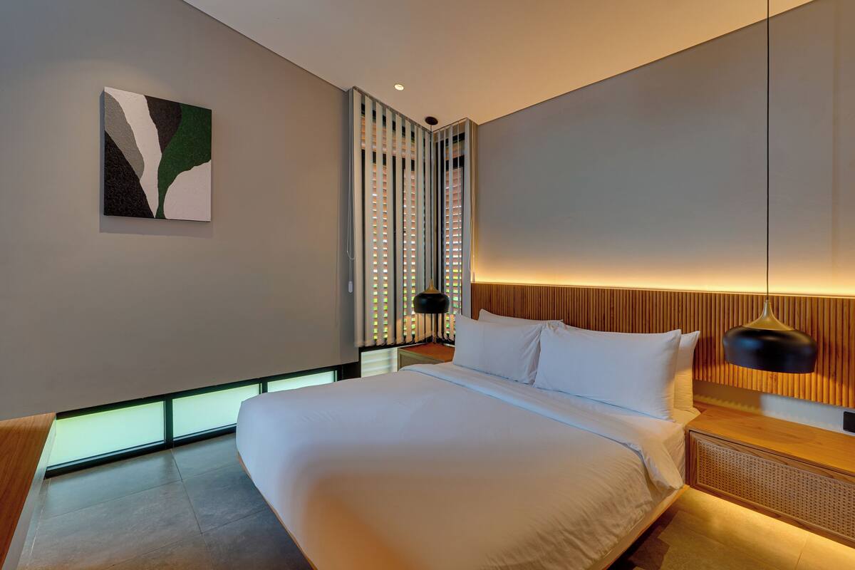 A modern hotel room with a large bed, a bedside table, a wall-mounted lamp, and a window with blinds.