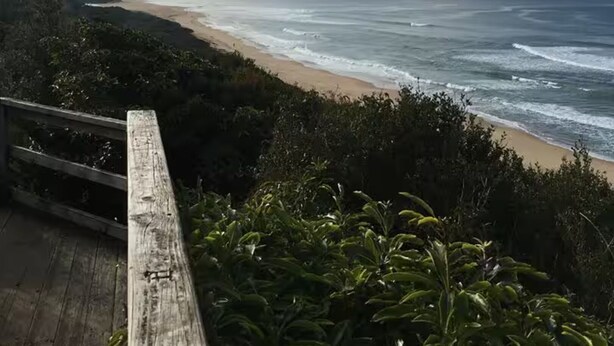 Une terrasse en bois donnant sur une plage, avec des vagues de l’océan et un ciel nuageux.