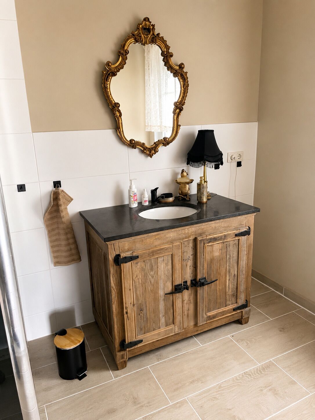 Een badkamer met een houten wastafelmeubel, een spiegel met een sierlijk omlijsting, een zwarte lamp en een handdoekrek.