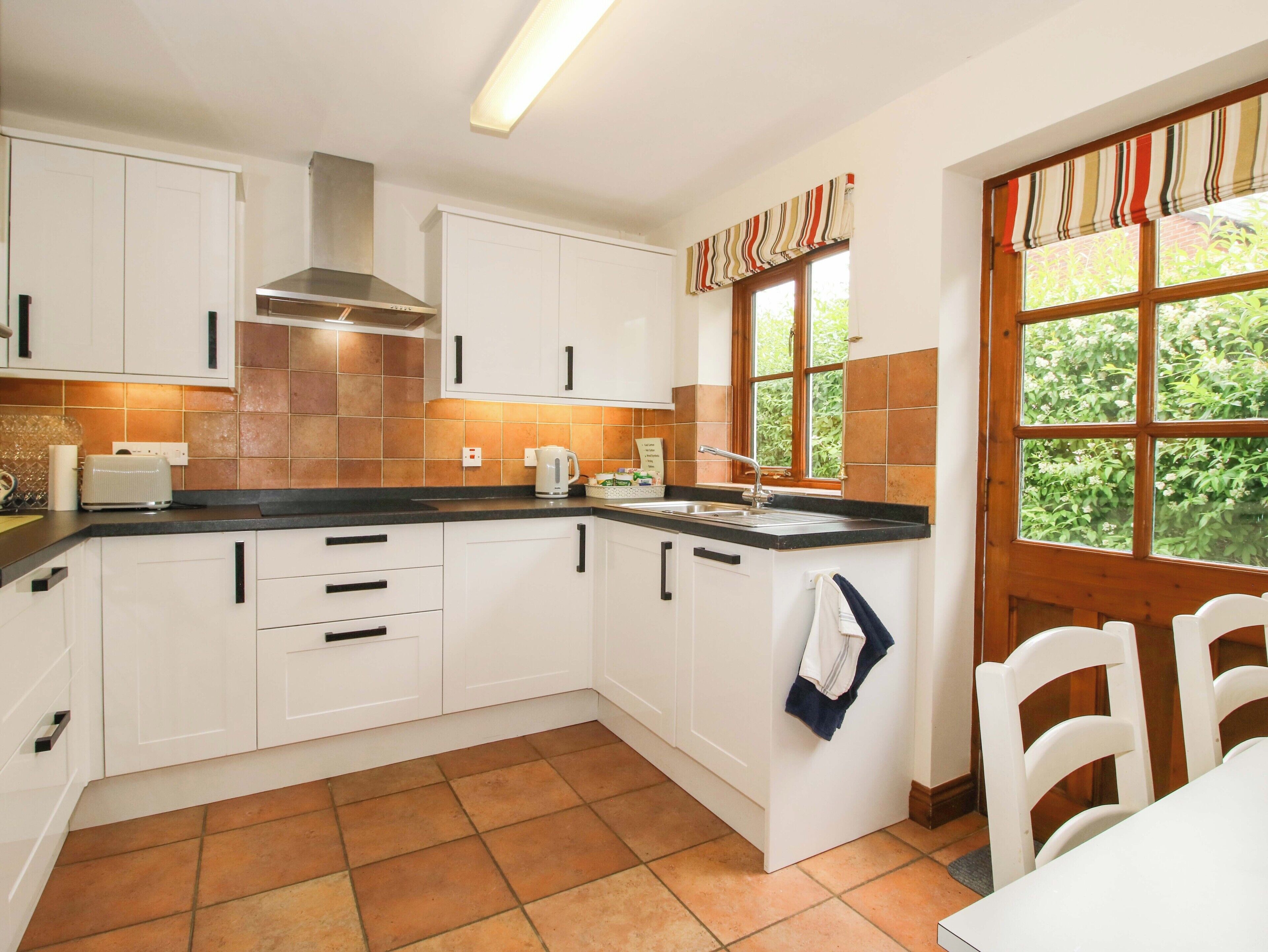 A kitchen with white cabinets, a black countertop, a stainless steel range hood, a window with a view of greenery, and a tiled floor.