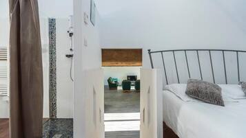 Une chambre avec un lit, un rideau de douche, une salle de bain avec un mur carrelé et une vue sur un espace de vie comprenant un téléviseur et des fauteuils.