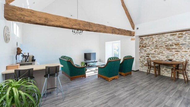 A spacious living area with a high ceiling, wooden beams, a stone wall, a dining table, and green upholstered chairs.
