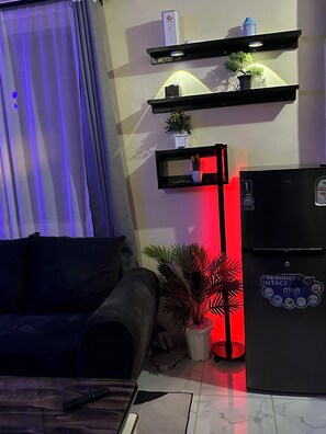 Ein Wohnzimmer mit einer Couch, einem schwarzen Kühlschrank, einer Topfpflanze und einem an der Wand befestigten Regal mit einer kleinen Pflanze und einer Wasserflasche.