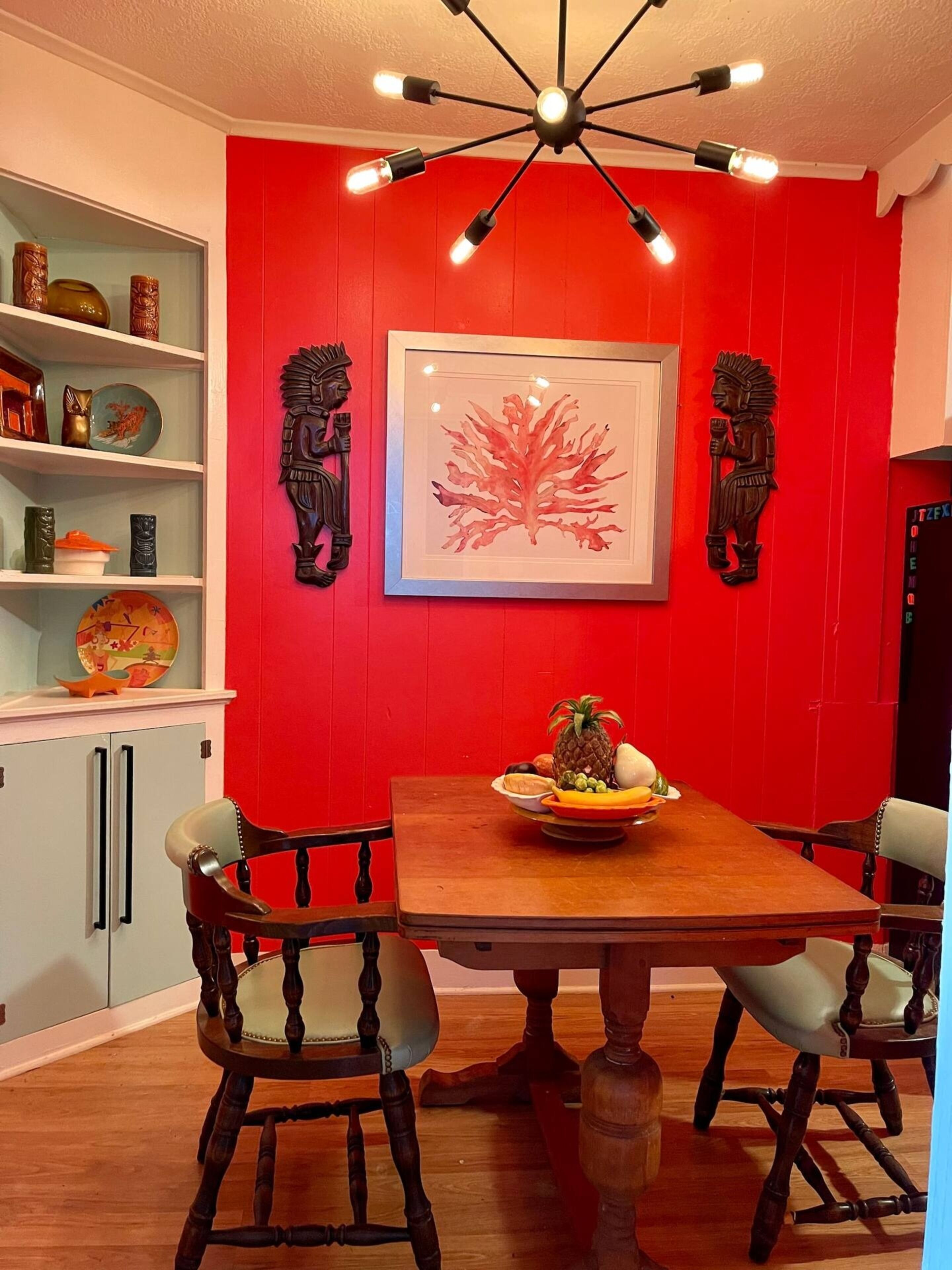 A dining area with a red accent wall, a wooden table, and chairs. There is a chandelier above the table, and a framed artwork is displayed on the wall.