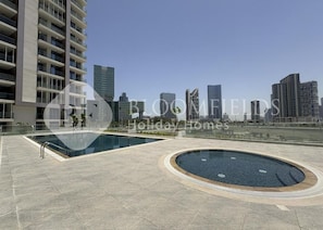 A modern residential building with a rooftop pool area, featuring a large rectangular pool and a circular hot tub, set against a city skyline. - Bloomfields 1BR Al Maryah Vista  (Abu Dhabi)