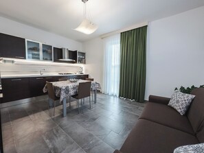 Eine moderne Küche mit dunklen Schränken, einem Esstisch mit Stühlen, einer Couch mit gemusterten Kissen und einem großen Fenster mit grünen Vorhängen.