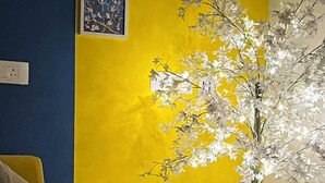 Ein Zimmer mit gelber Wand, einem braunen Sofa, einem runden Tisch mit Kerze und einer Topfpflanze mit weißen Blüten.